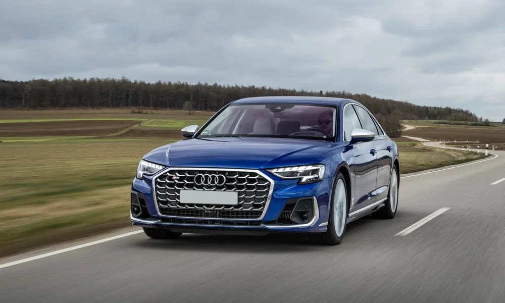 Фото Audi S8 (2024) — вид спереди, фото 7. Синий кузов, строгая симметрия оптики — базовый ракурс знакомства с дизайном. Рисунок ходовых огней — подпись модели, по нему её узнают в потоке Audi S8 (2024) — вид спереди, фото 7