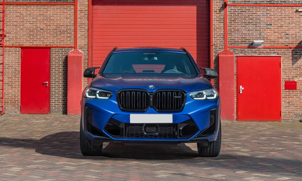 Фото BMW X3 M (2024) — вид спереди, фото 6. Кузов в синем цвете: рисунок оптики и решётки задаёт узнаваемое «лицо» модели. Рисунок ходовых огней — подпись модели, по нему её узнают в потоке BMW X3 M (2024) — вид спереди, фото 6