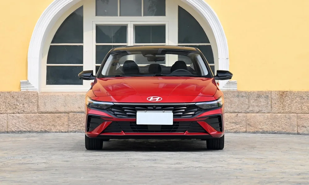 Фото Hyundai Elantra (2024) — вид спереди, фото 4. Кузов в красном цвете: рисунок оптики и решётки задаёт узнаваемое «лицо» модели. Рисунок ходовых огней — подпись модели, по нему её узнают в потоке Hyundai Elantra (2024) — вид спереди, фото 4