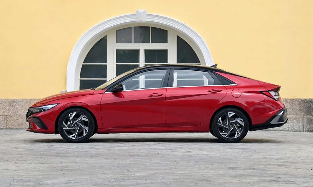 Фото Hyundai Elantra (2024) — вид сбоку, фото 2. Сбоку читаются линия остекления и характер выштамповок на красном кузове. Хромированные или чёрные молдинги окон отличают комплектации Hyundai Elantra (2024) — вид сбоку, фото 2