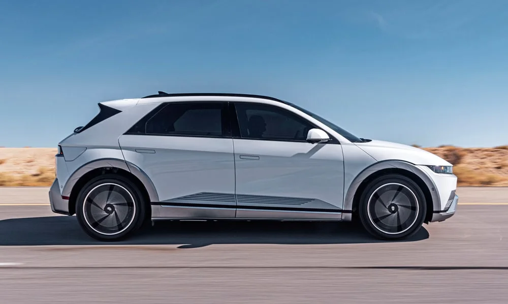 Фото Hyundai IONIQ 5 (2024) — вид сбоку, фото 3. Сбоку читаются линия остекления и характер выштамповок на белом кузове. Хромированные или чёрные молдинги окон отличают комплектации Hyundai IONIQ 5 (2024) — вид сбоку, фото 3