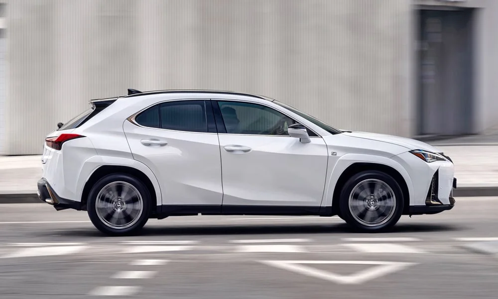 Фото Lexus UX 200 (2024) — вид сбоку, фото 2. Сбоку читаются линия остекления и характер выштамповок на белом кузове. Хромированные или чёрные молдинги окон отличают комплектации Lexus UX 200 (2024) — вид сбоку, фото 2