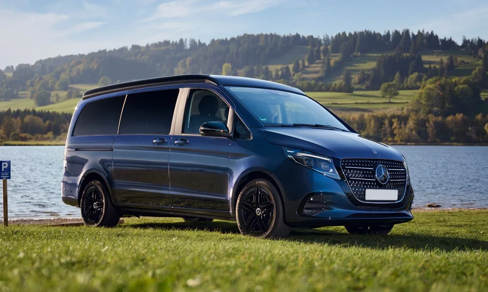 Фото Mercedes-Benz V-Class (2024) — вид спереди, фото 9. Синий кузов, строгая симметрия оптики — базовый ракурс знакомства с дизайном. Рисунок ходовых огней — подпись модели, по нему её узнают в потоке Mercedes-Benz V-Class (2024) — вид спереди, фото 9