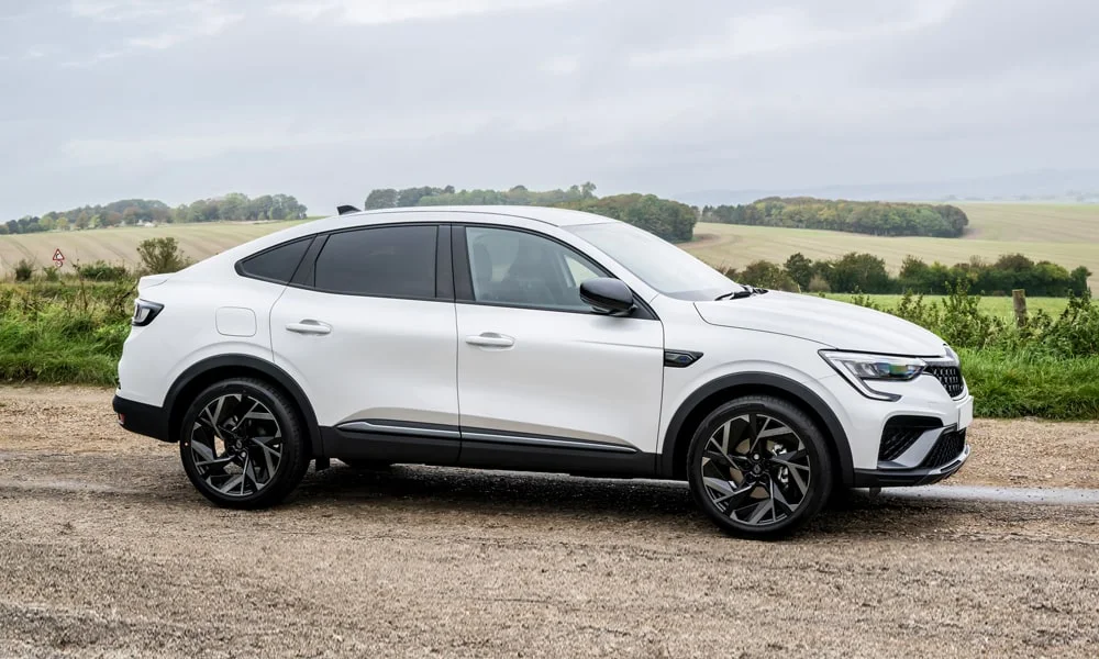 Фото Renault Arkana (2024) — вид сбоку, фото 5. Сбоку читаются линия остекления и характер выштамповок на белом кузове. Хромированные или чёрные молдинги окон отличают комплектации Renault Arkana (2024) — вид сбоку, фото 5