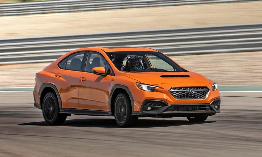 Фото Subaru WRX STI (2024) — вид спереди, фото 7. Оранжевый кузов, строгая симметрия оптики — базовый ракурс знакомства с дизайном. Рисунок ходовых огней — подпись модели, по нему её узнают в потоке Subaru WRX STI (2024) — вид спереди, фото 7