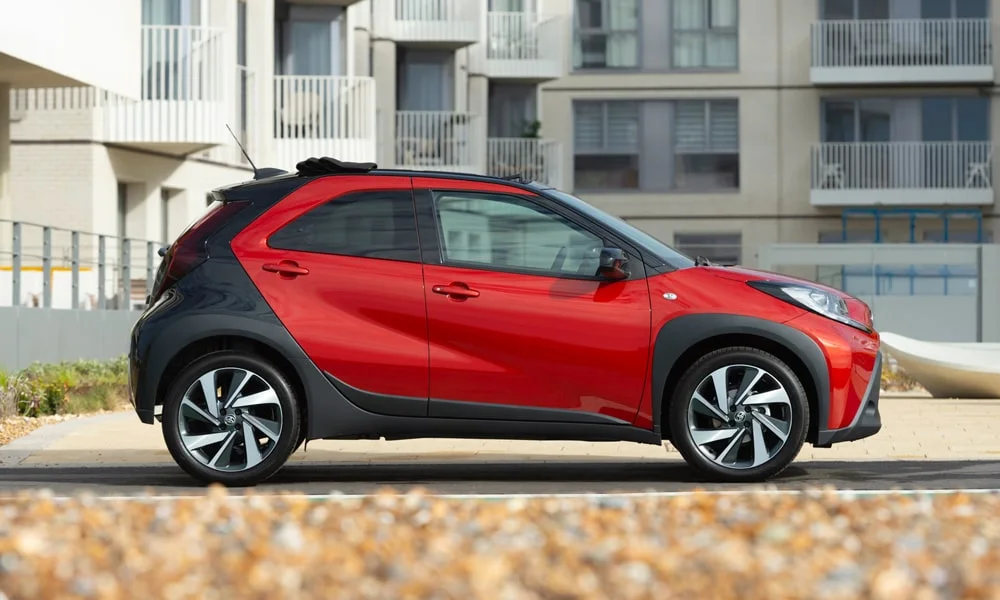 Фото Toyota Aygo X — вид сбоку, фото 4. Красный кузов в профиль — пропорции, высота посадки и дизайн дисков. Хромированные или чёрные молдинги окон отличают комплектации Toyota Aygo X — вид сбоку, фото 4